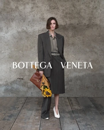 Model wearing dark blazer and skirt holding a leather bag with sunflowers, set against a rustic stone wall.
