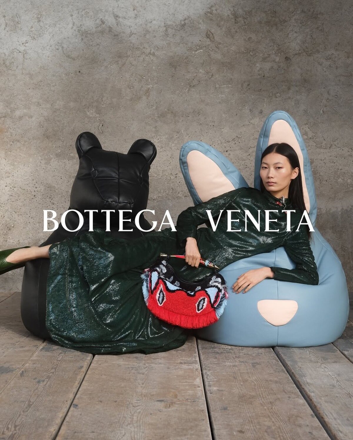 Model wearing a dark green dress reclining against a blue rabbit-eared beanbag chair next to a black bear beanbag chair.