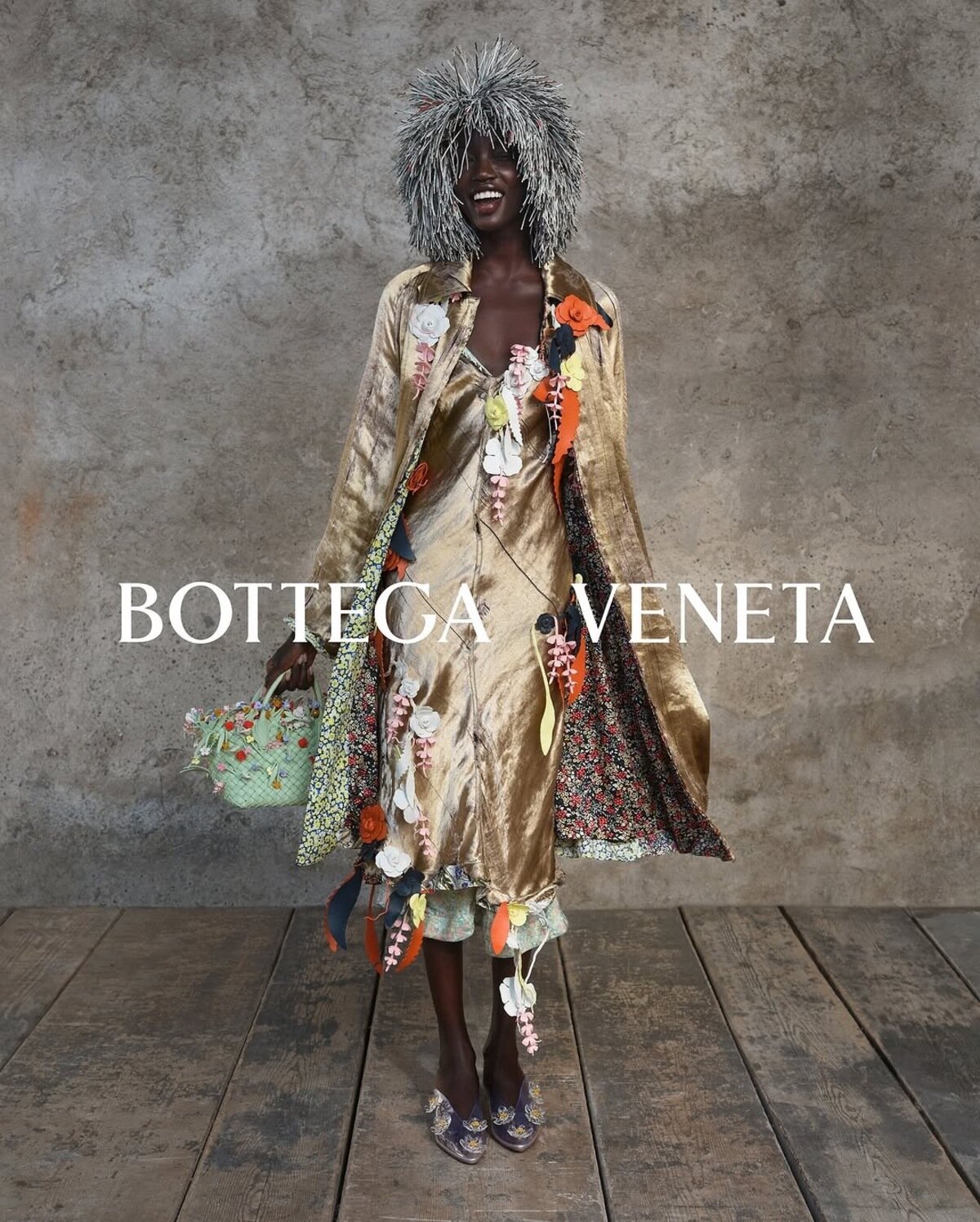 Model wearing a metallic gold coat and matching dress adorned with floral appliqués, holding an embellished woven handbag against a concrete wall