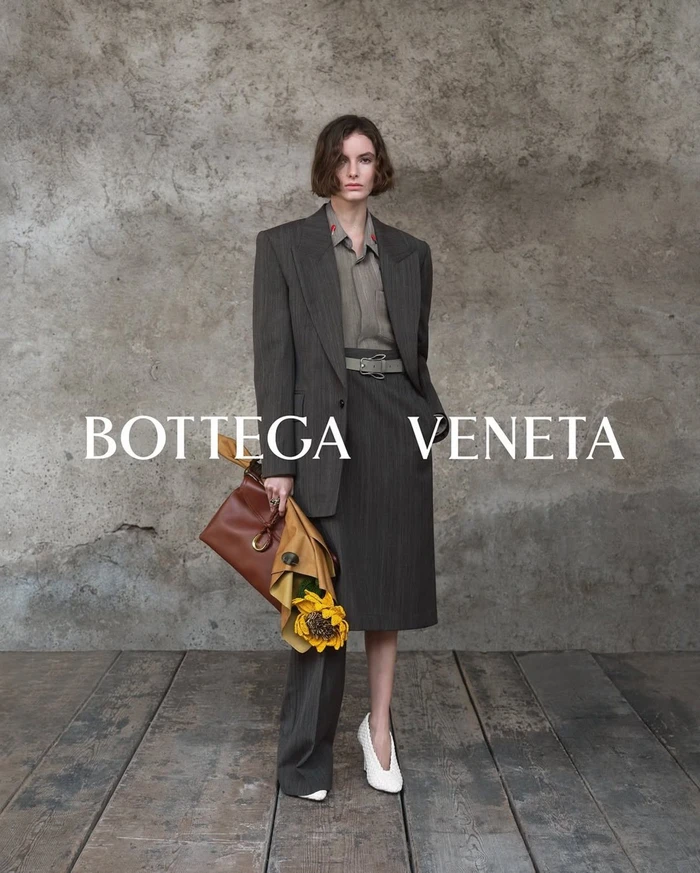Model wearing dark blazer and skirt holding a leather bag with sunflowers, set against a rustic stone wall.