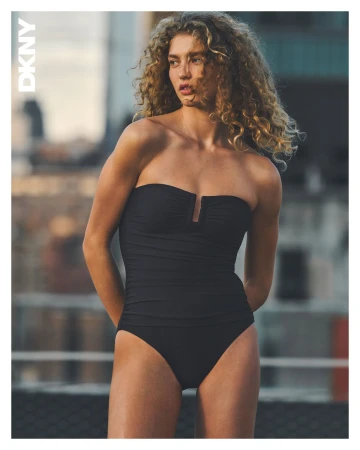 Woman with curly blonde hair wearing a black strapless swimsuit standing against a blurred urban background