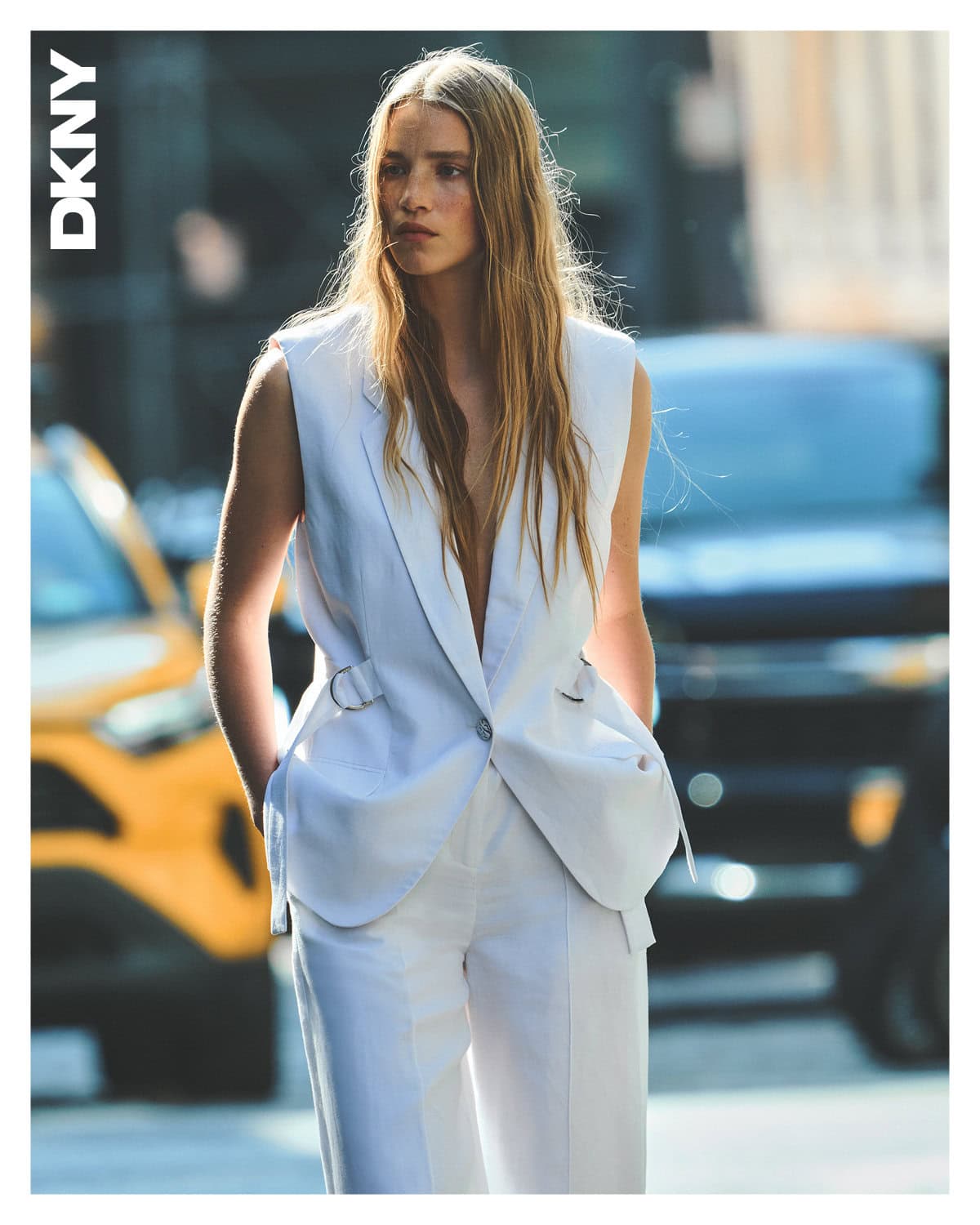 Model with long blonde hair wearing a white sleeveless vest and trousers on an urban street.