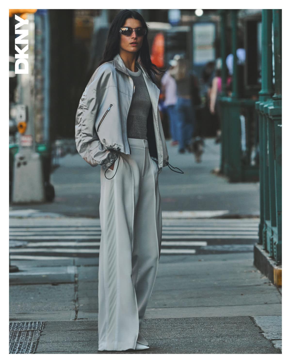 Model wearing a light grey bomber jacket and wide-leg trousers standing on a city street for DKNY Spring 2025 campaign.