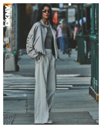 Model wearing a light grey bomber jacket and wide-leg trousers standing on a city street for DKNY Spring 2025 campaign.