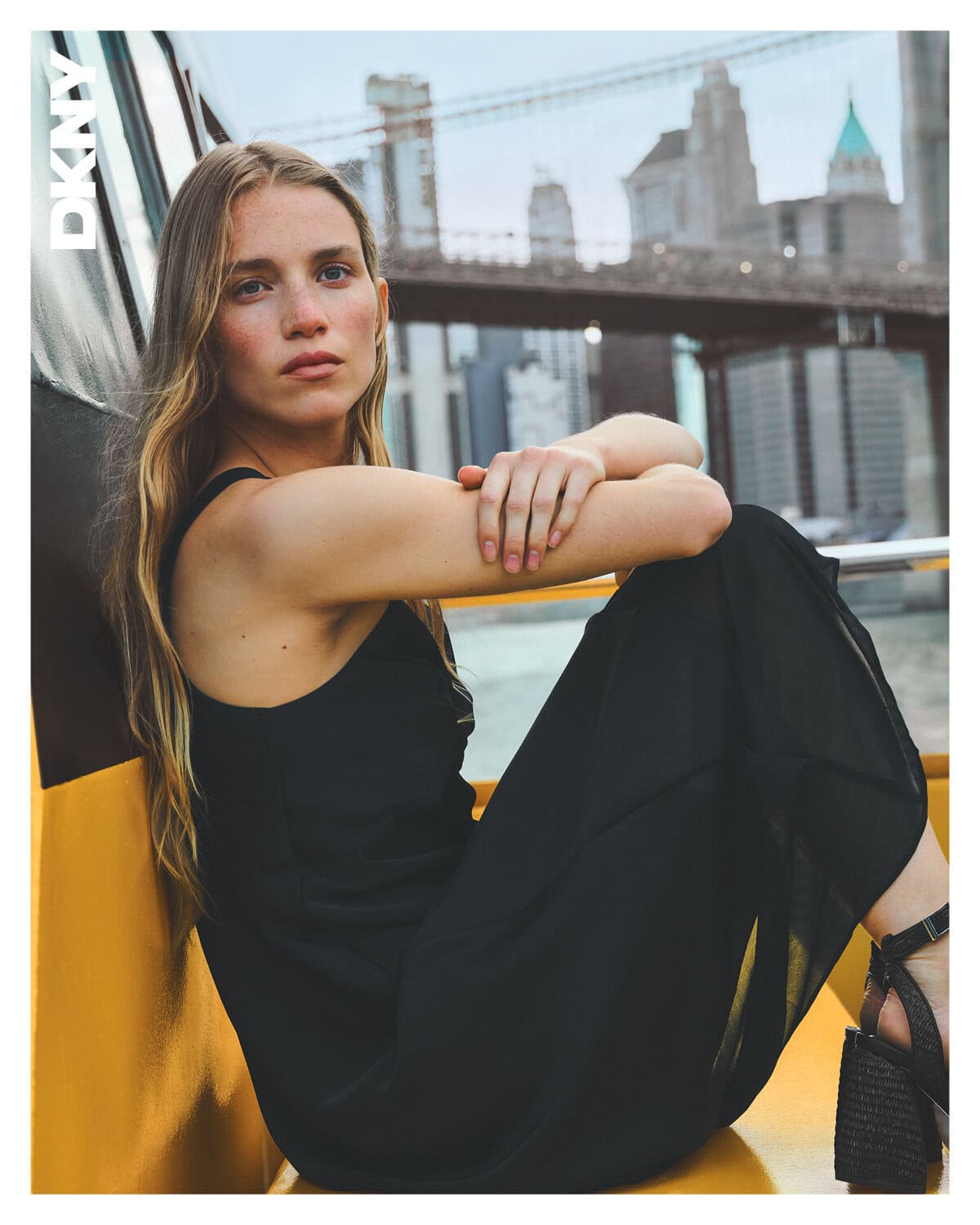 Woman in a black dress sitting on a yellow structure with a bridge and city skyline visible in the background.