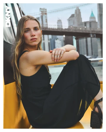Woman in a black dress sitting on a yellow structure with a bridge and city skyline visible in the background.