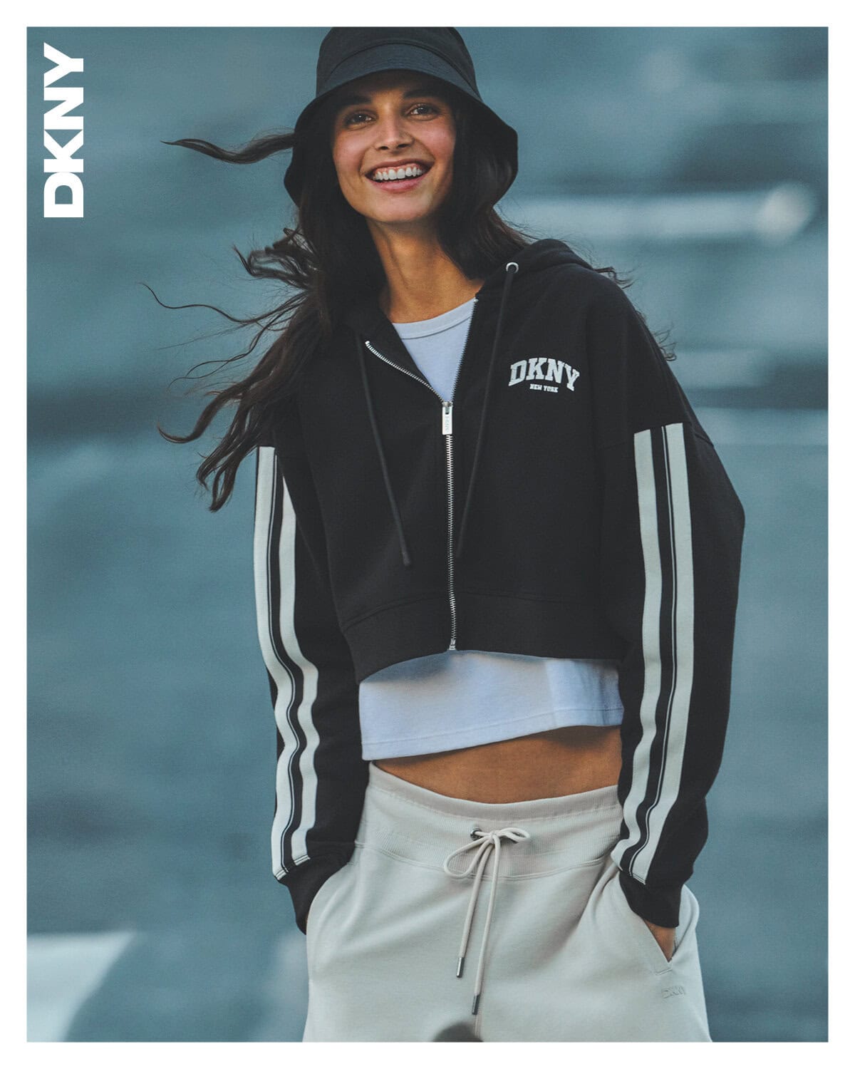 Smiling female model wearing a black zip hoodie, white t-shirt, sweatpants, and bucket hat against an outdoor backdrop