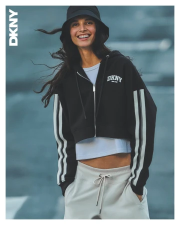 Smiling female model wearing a black zip hoodie, white t-shirt, sweatpants, and bucket hat against an outdoor backdrop
