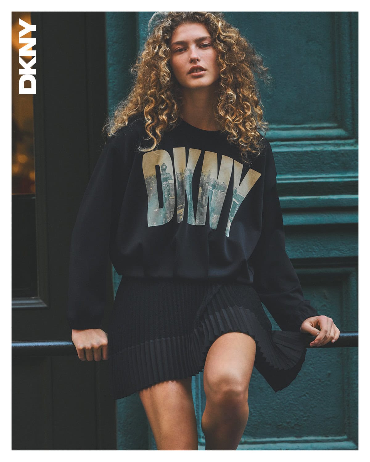 Model with long curly hair wearing a black sweatshirt with DKNY logo and pleated skirt against a dark green wooden door.
