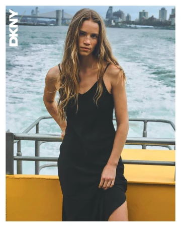 Model in a black slip dress standing on a boat with water and city skyline in background