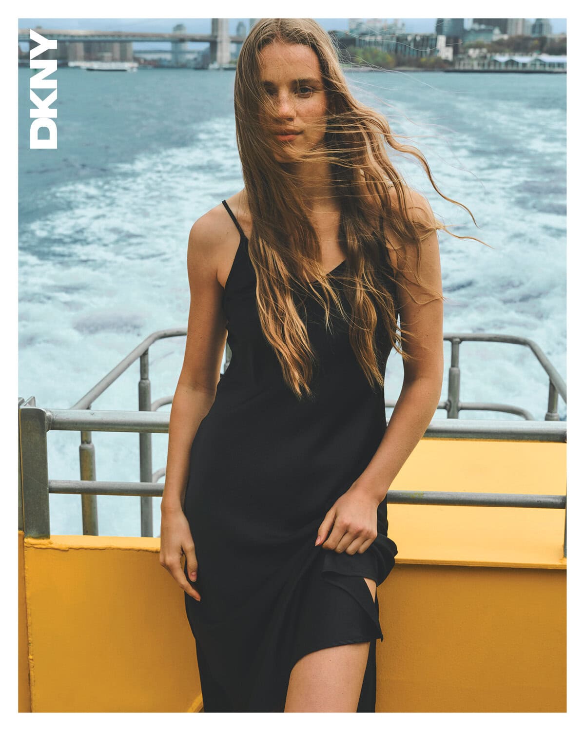 Model in black slip dress standing on a boat deck with water and distant bridge in the background for DKNY Spring 2025 ad campaign