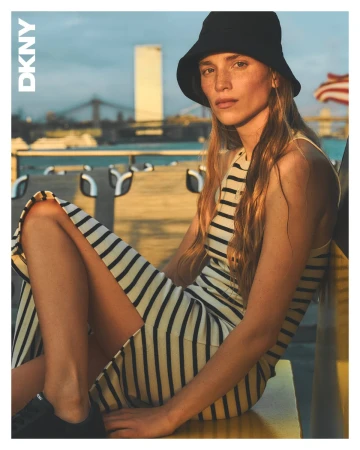 Model in a black bucket hat and striped dress seated outdoors near water with a bridge and skyline background.