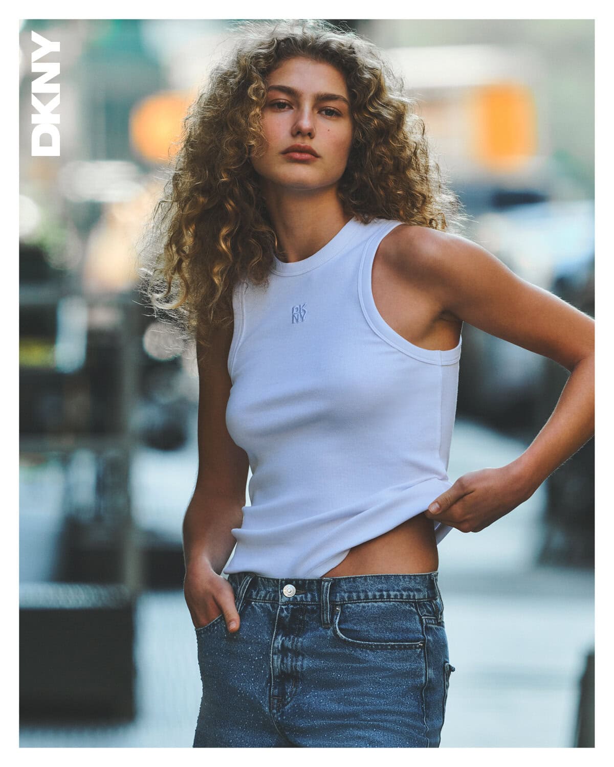 Model with curly hair wearing a white tank top and blue jeans against a blurred urban street background