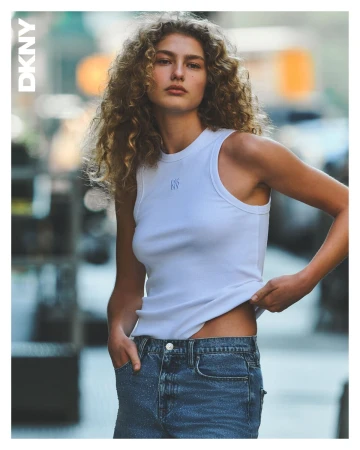 Model with curly hair wearing a white tank top and blue jeans against a blurred urban street background