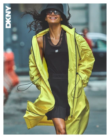 Smiling model in a bright yellow windbreaker, black tank top, skirt, bucket hat, and sunglasses on an urban street.