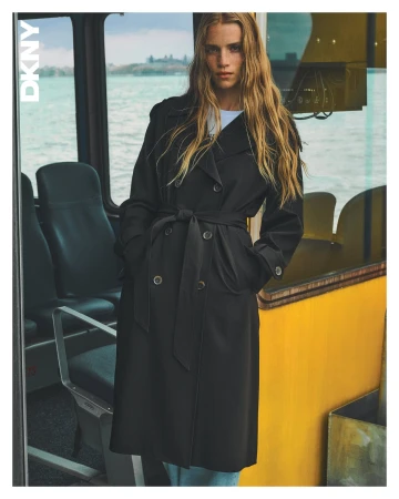 Model with long blonde hair wearing a belted black trench coat inside a ferry cabin with water visible through the windows