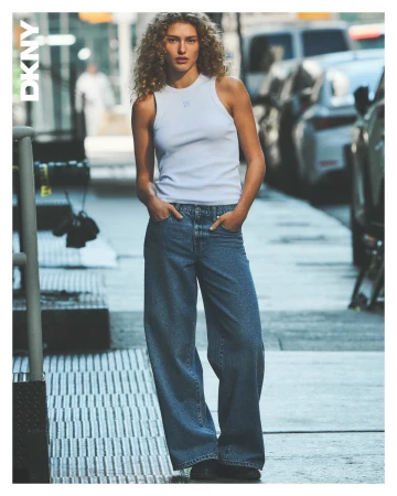 Model with curly blonde hair wearing a white tank top and blue jeans standing on a city street