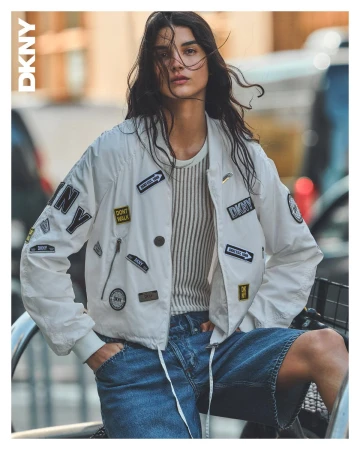 Model wearing a white bomber jacket with patches and blue denim shorts outdoors with soft natural lighting.