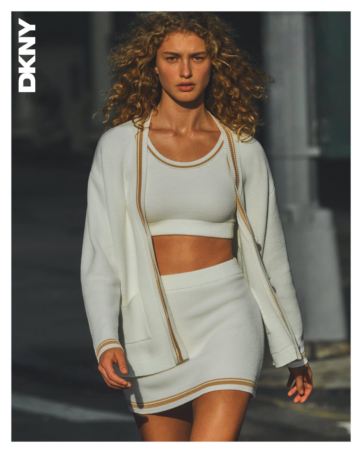 Model with curly blonde hair wearing a white knit cardigan, crop top, and skirt on an urban street.