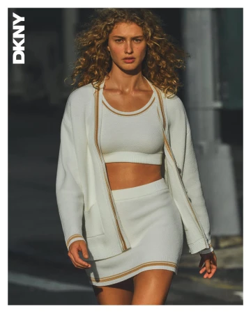 Model with curly blonde hair wearing a white knit cardigan, crop top, and skirt on an urban street.