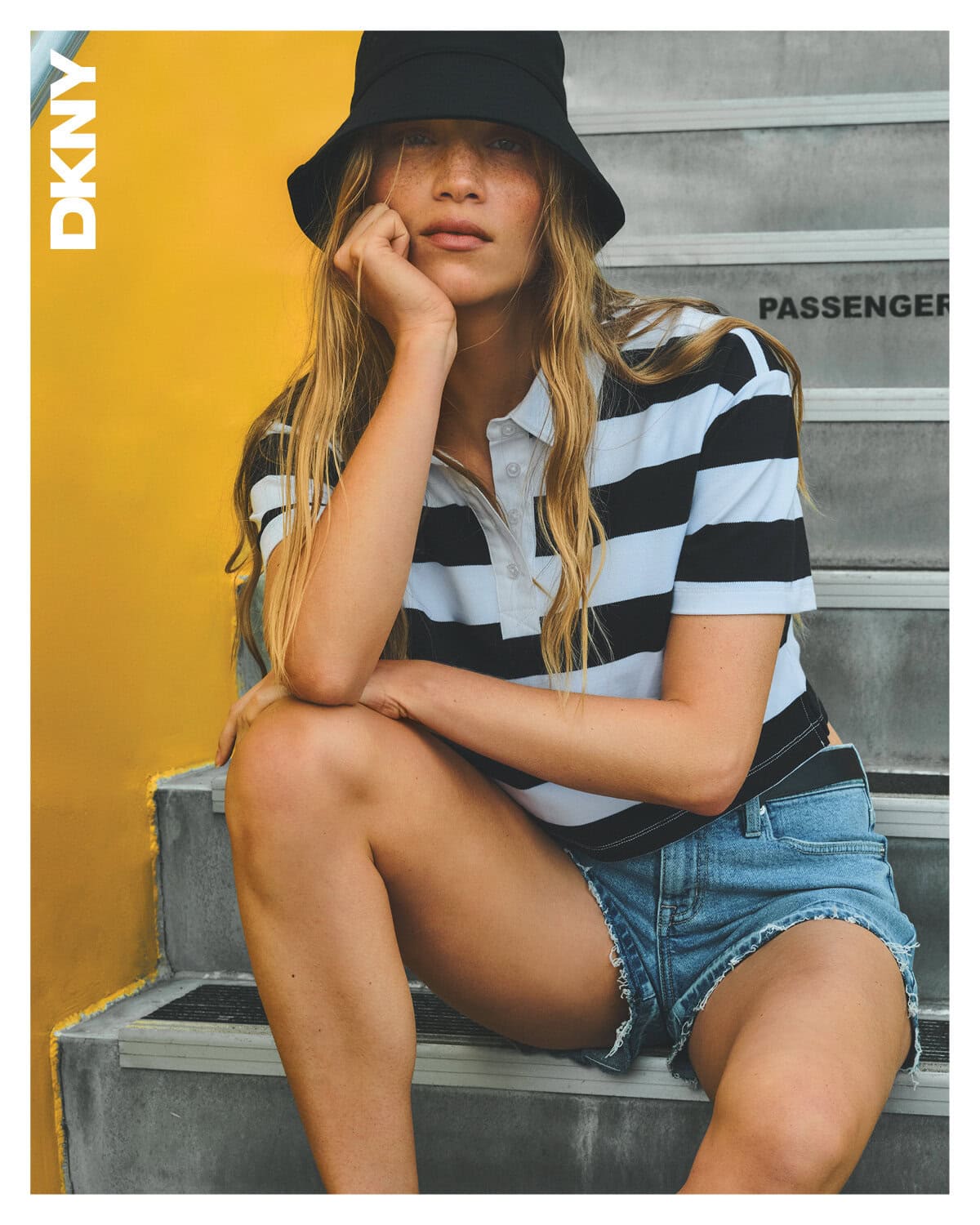 Model wearing a black bucket hat, striped polo shirt, and denim shorts sitting on outdoor concrete steps next to a yellow wall.
