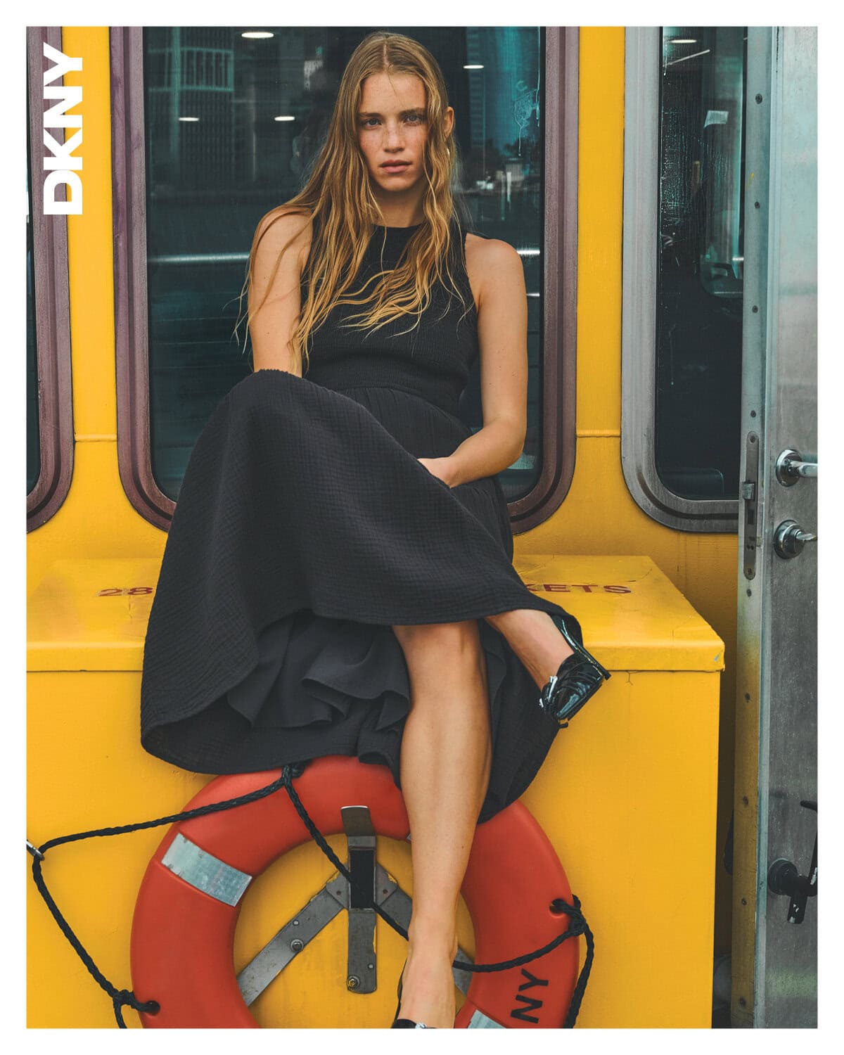 Model wearing a black dress seated on a bright yellow structure with a red lifebuoy underneath