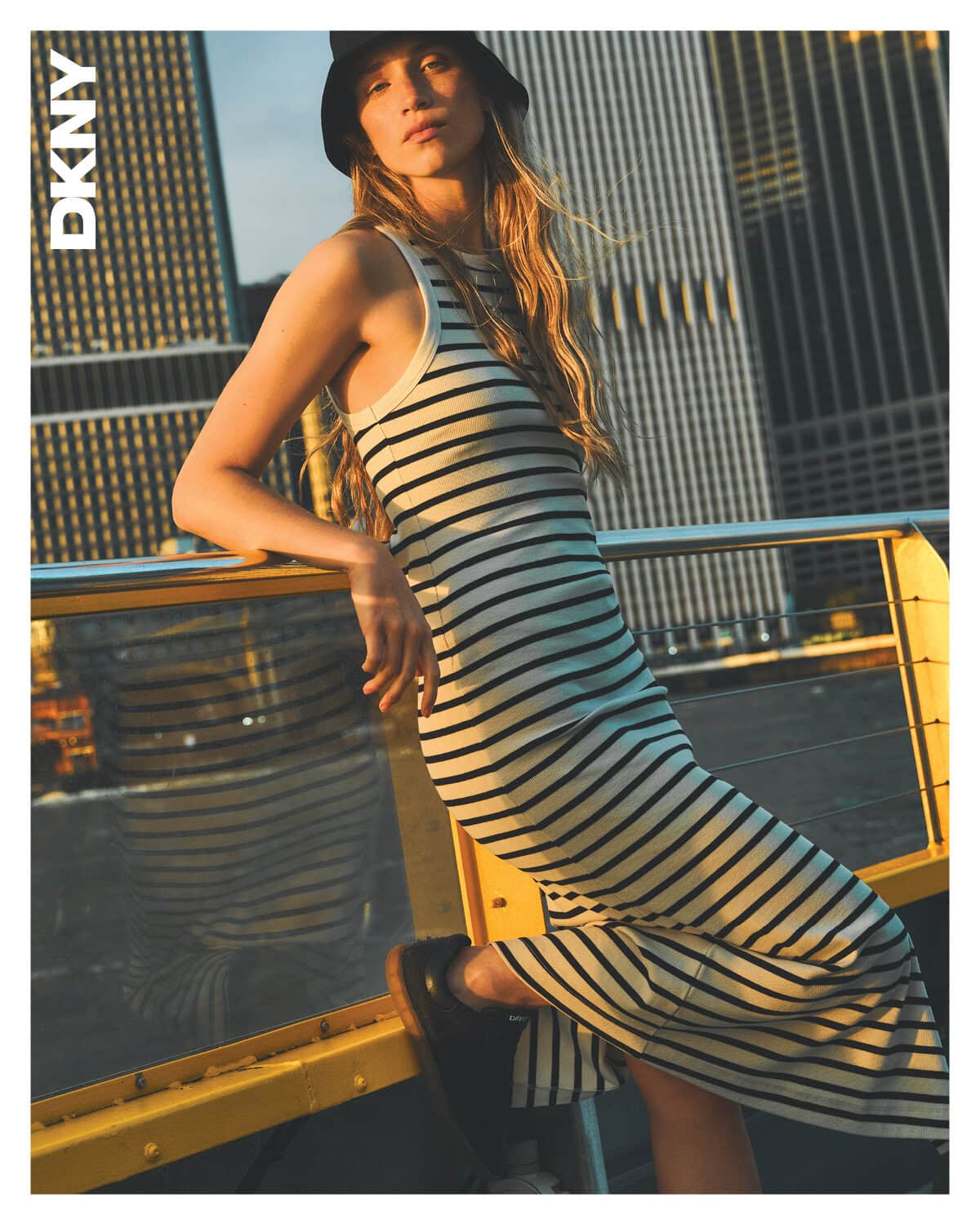 Model wearing a striped dress and bucket hat leaning against a yellow railing outdoors with skyscrapers behind