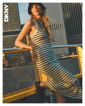 Model wearing a striped dress and bucket hat leaning against a yellow railing outdoors with skyscrapers behind