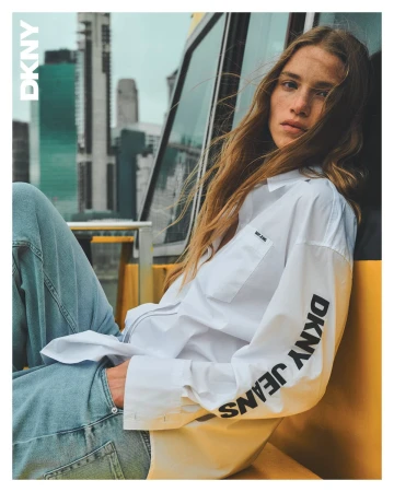Model with long brown hair wearing a white DKNY long-sleeve shirt and blue jeans, leaning against a yellow structure with city background.