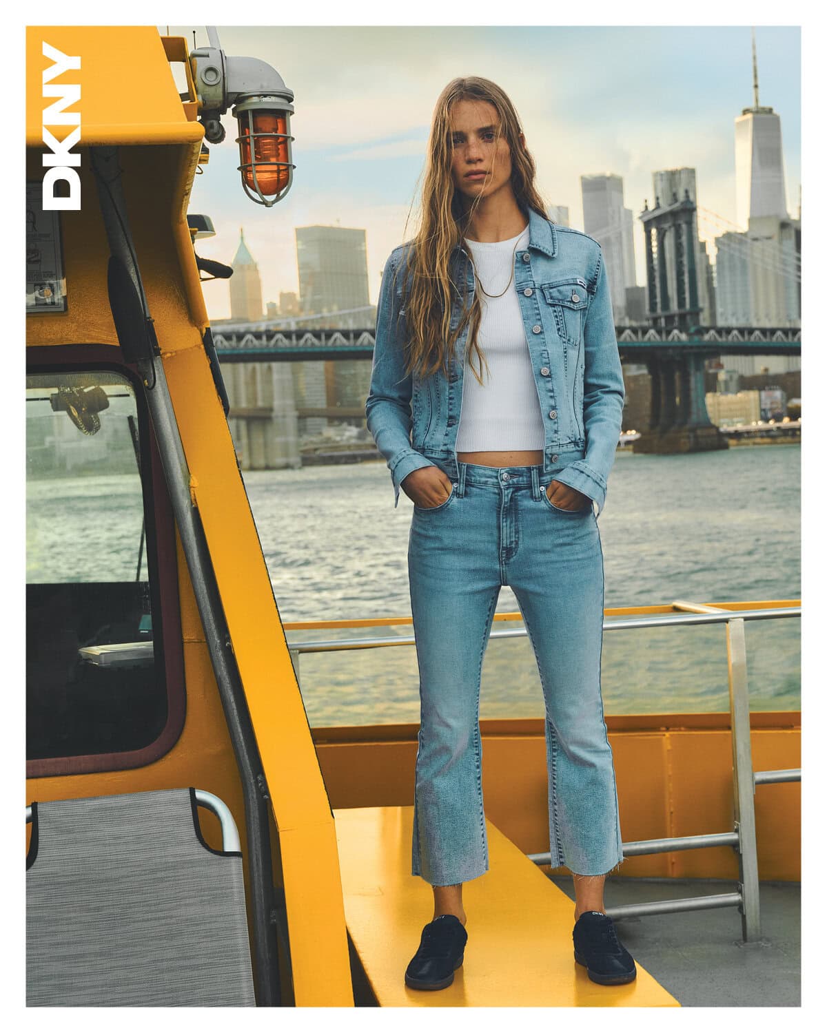 Model wearing a denim jacket and jeans standing on a yellow boat deck with a city skyline and bridge in background