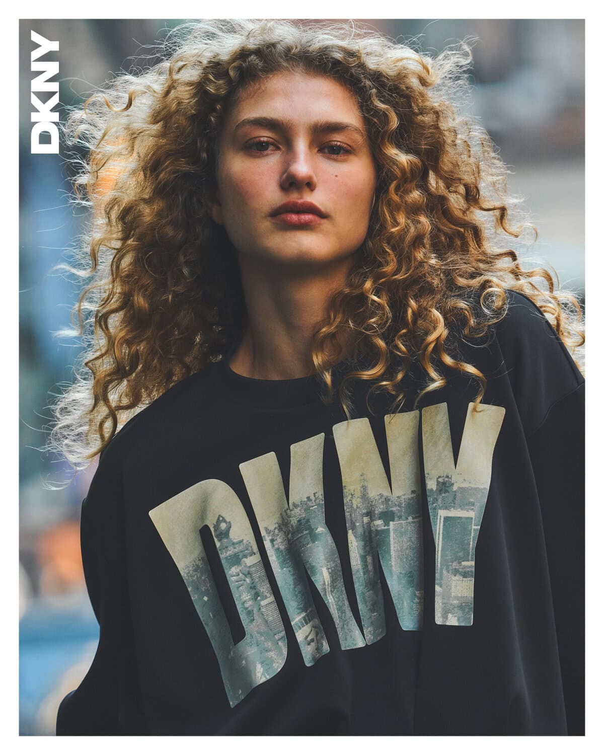 Model with long curly blonde hair wearing a black sweatshirt with large DKNY logo lettering against an urban street background.