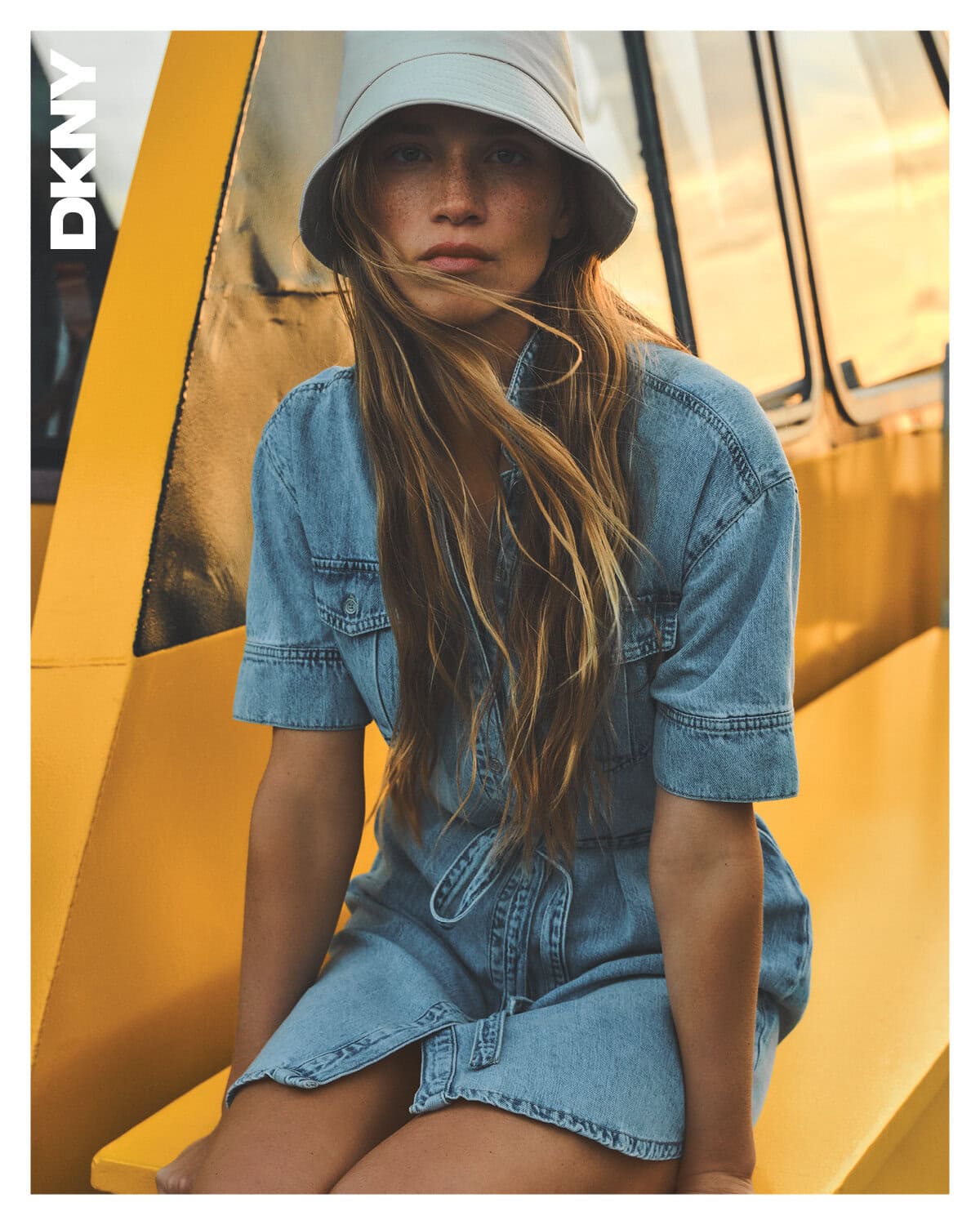 Model in denim dress and bucket hat seated against a yellow backdrop for DKNY Spring 2025 campaign