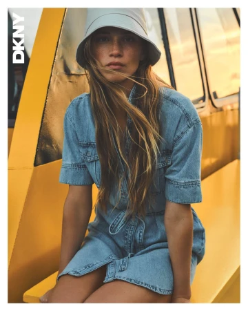 Model in denim dress and bucket hat seated against a yellow backdrop for DKNY Spring 2025 campaign