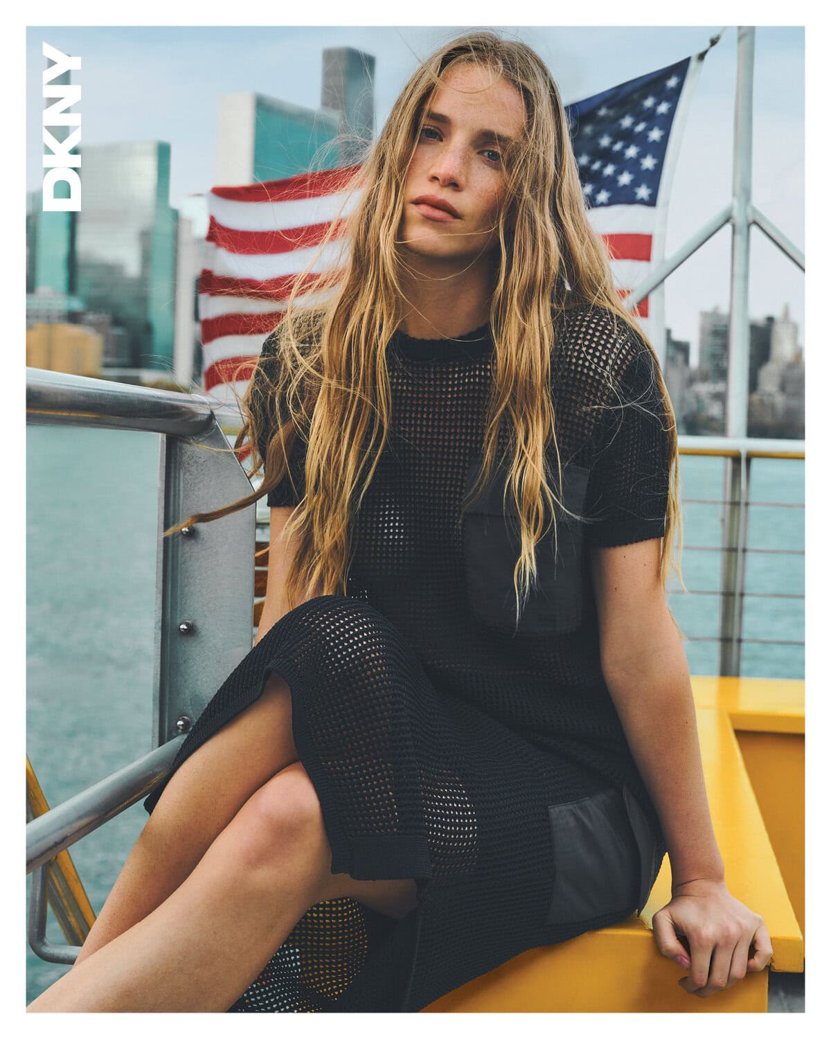 Model seated outdoors on a yellow ferry railing wearing a black mesh dress with an American flag and city skyline background.