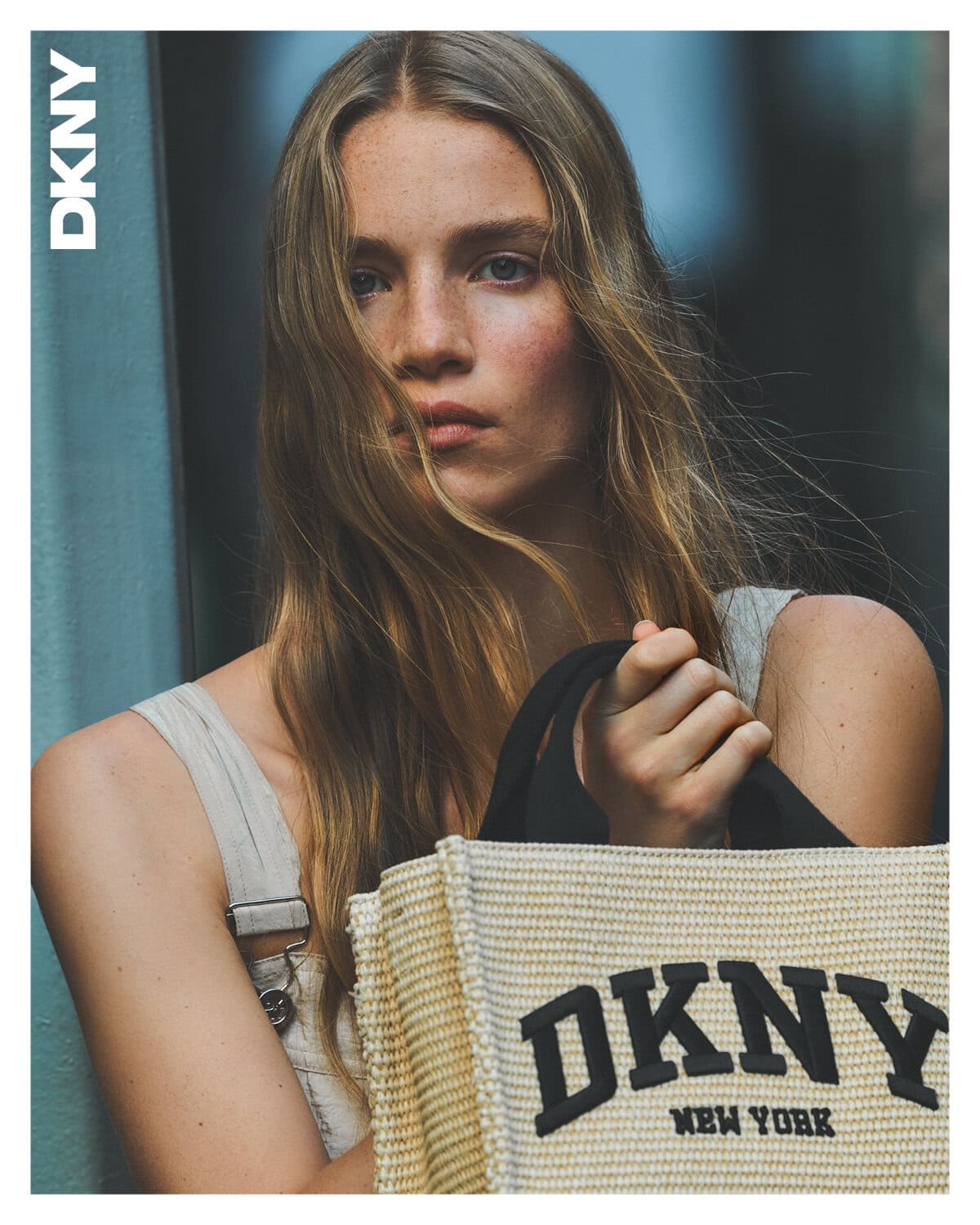 Model with long blonde hair holding a textured canvas tote bag with DKNY branding against an urban street background.