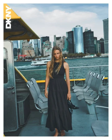 Model standing on a ferry deck in a black maxi dress holding shoes, with a New York City skyline background.