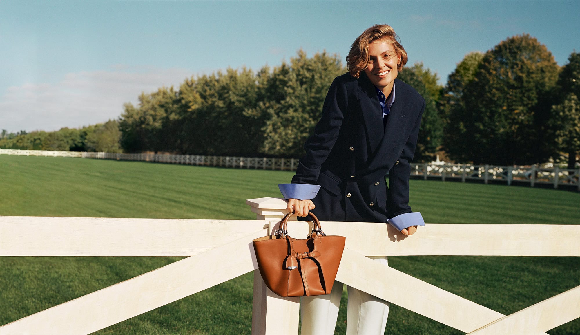 Woman in dark blazer leaning on a white fence holding a brown leather bag outdoors against a grassy field and trees