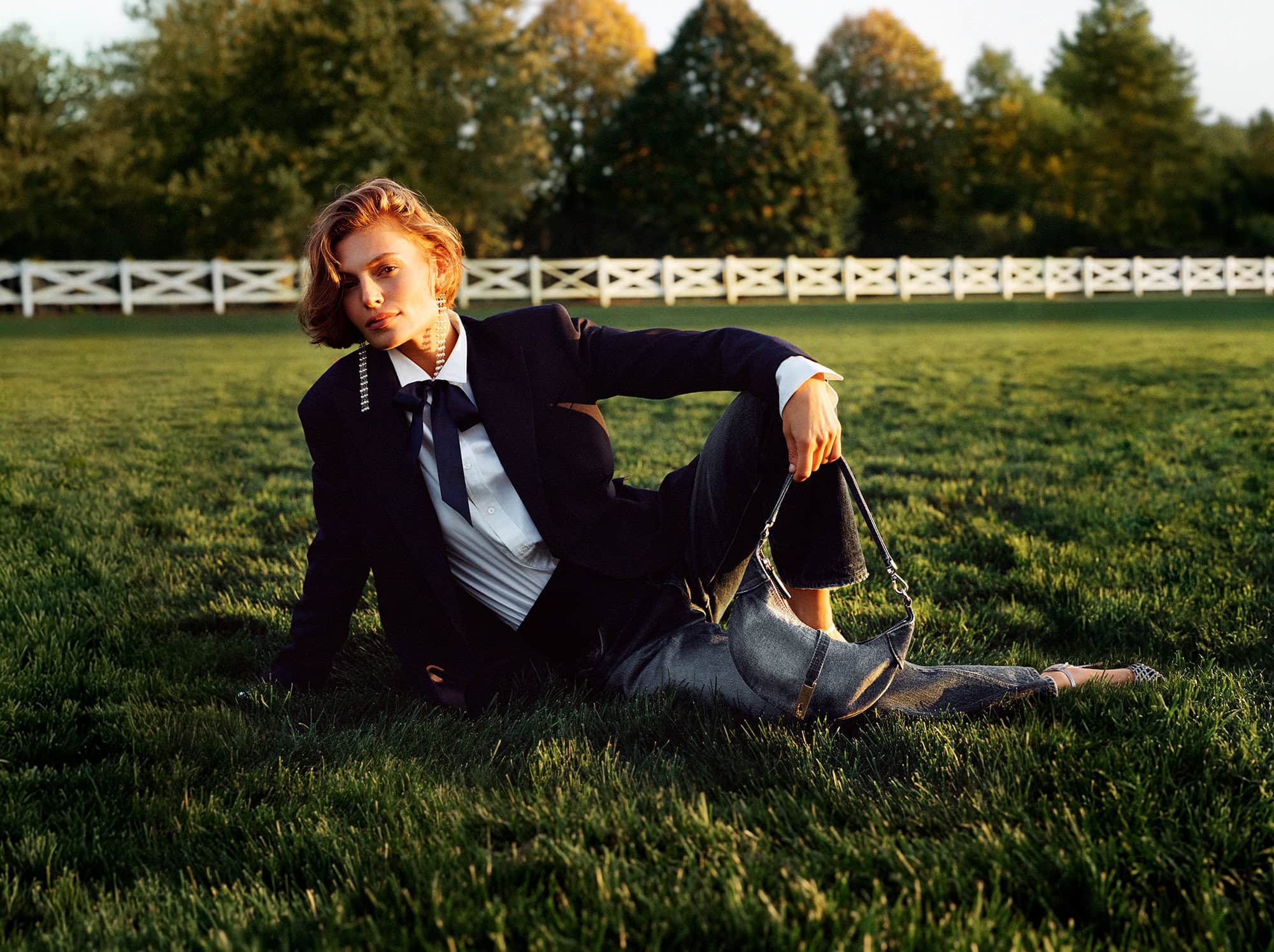 Model seated on green grass wearing a dark blazer, white shirt, and bow tie, holding a handbag, with a white fence behind.