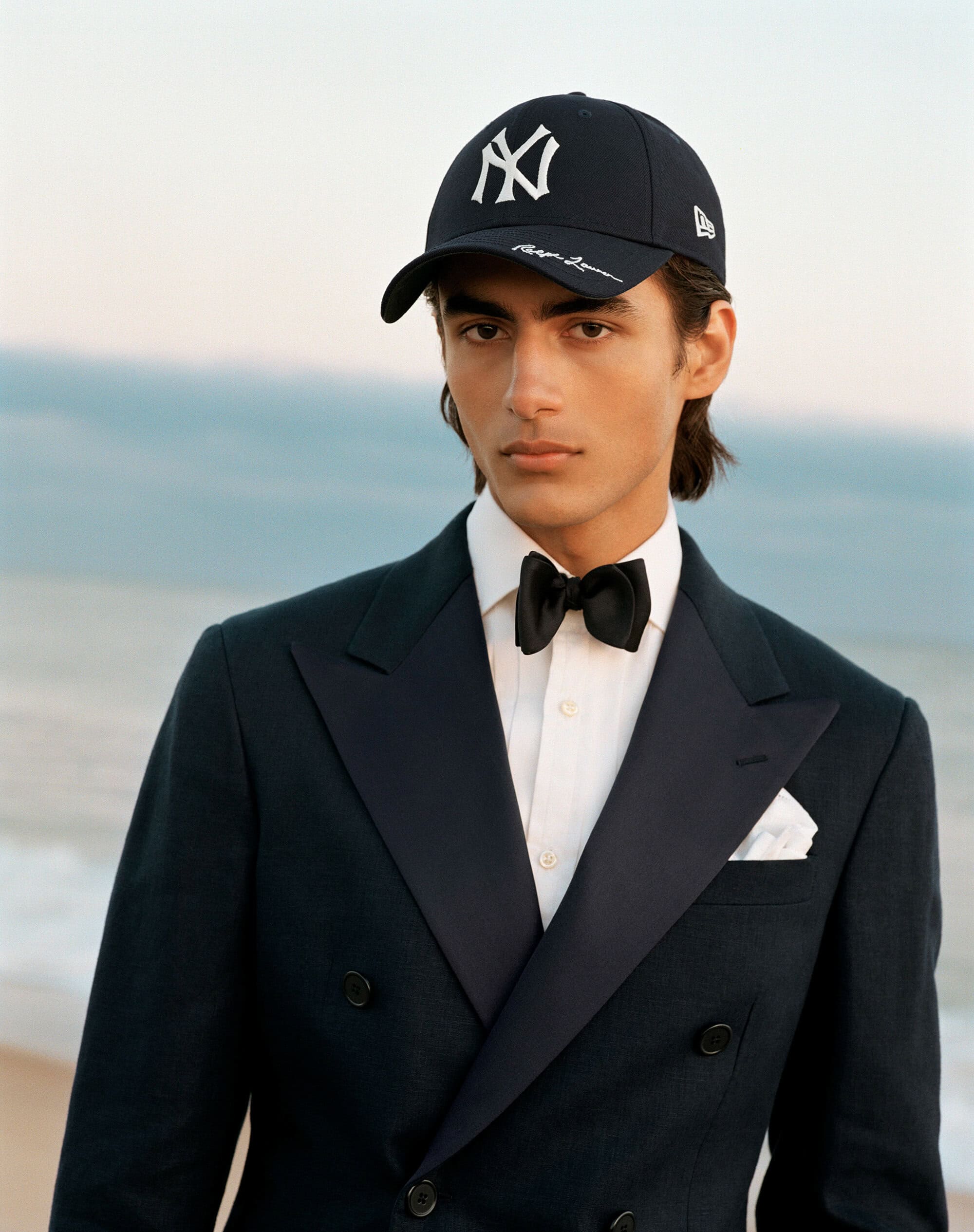 Young man wearing a dark tuxedo jacket, white shirt, black bow tie, and a baseball cap against a blurred coastal backdrop