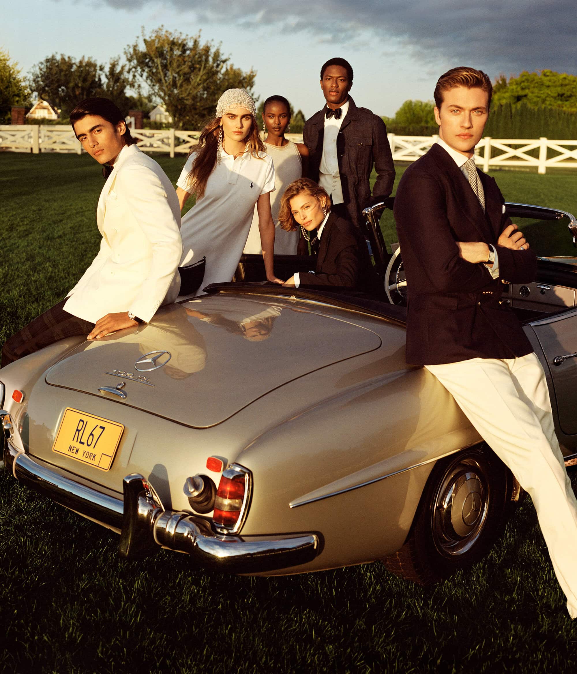 Group of six models posing around a vintage convertible car on a green lawn with a white fence background.