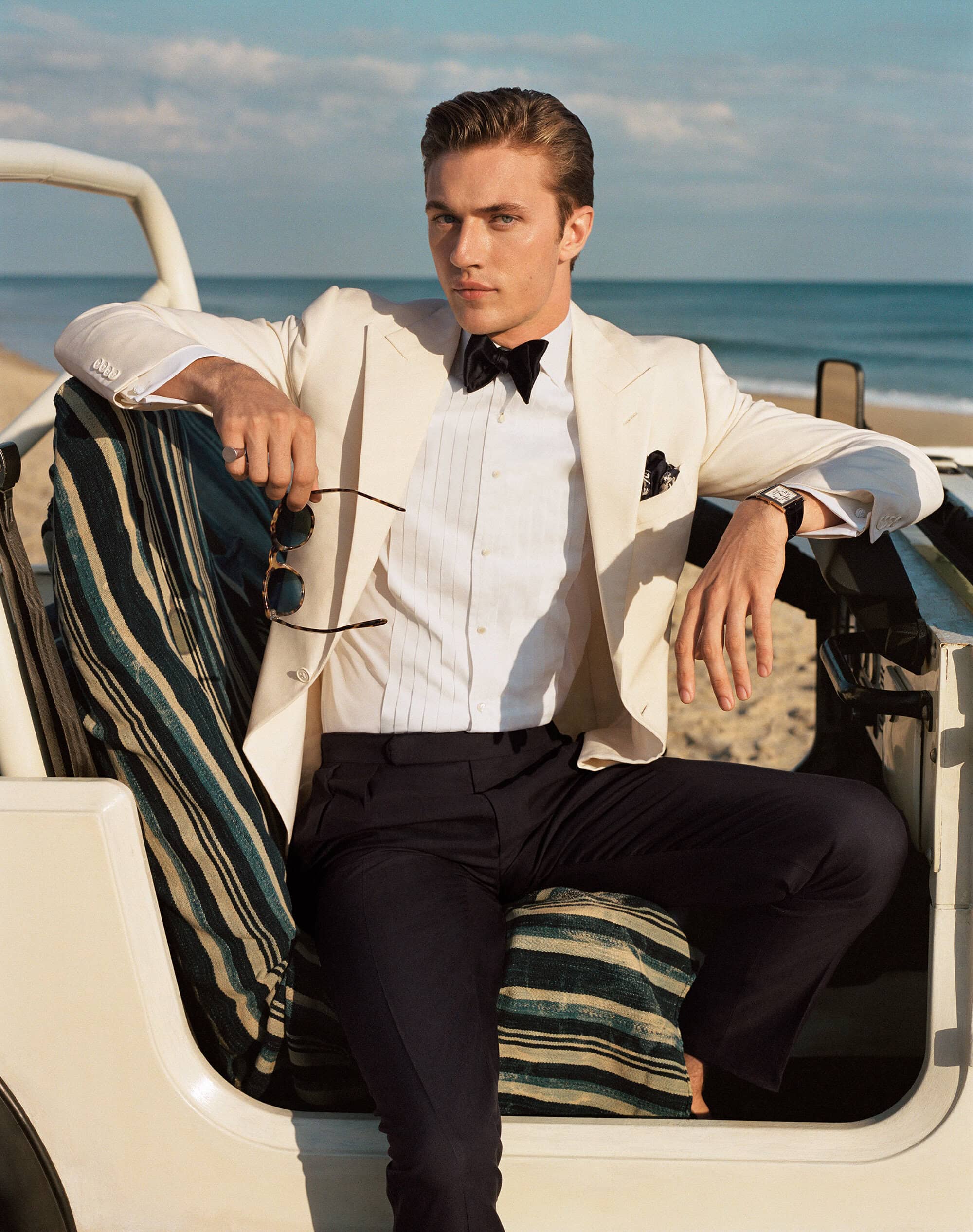 Man wearing ivory suit jacket and bow tie sitting in a vintage beach car with ocean background