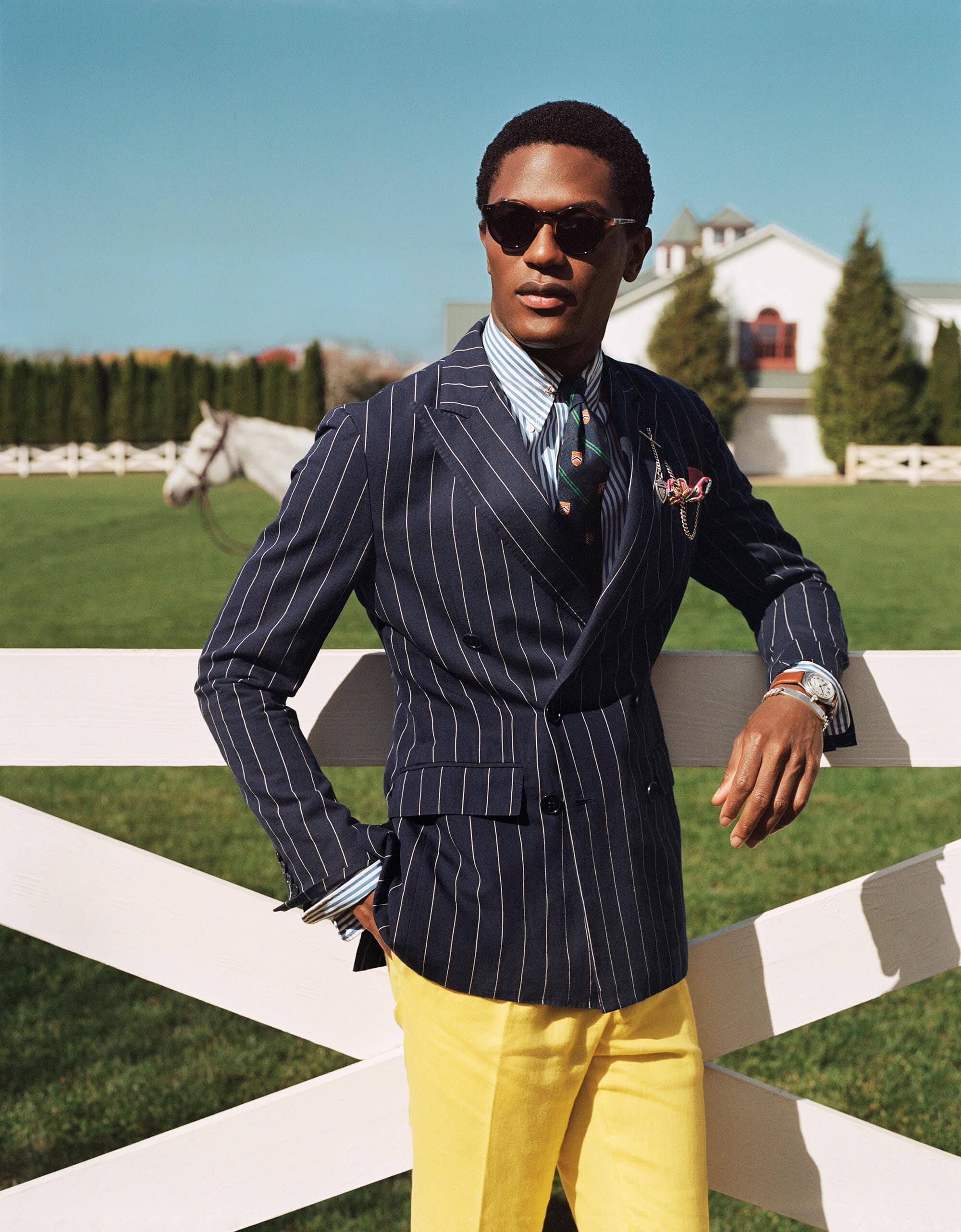 Man wearing a navy pinstripe suit jacket and yellow trousers leans against a white wooden fence outdoors in daylight.