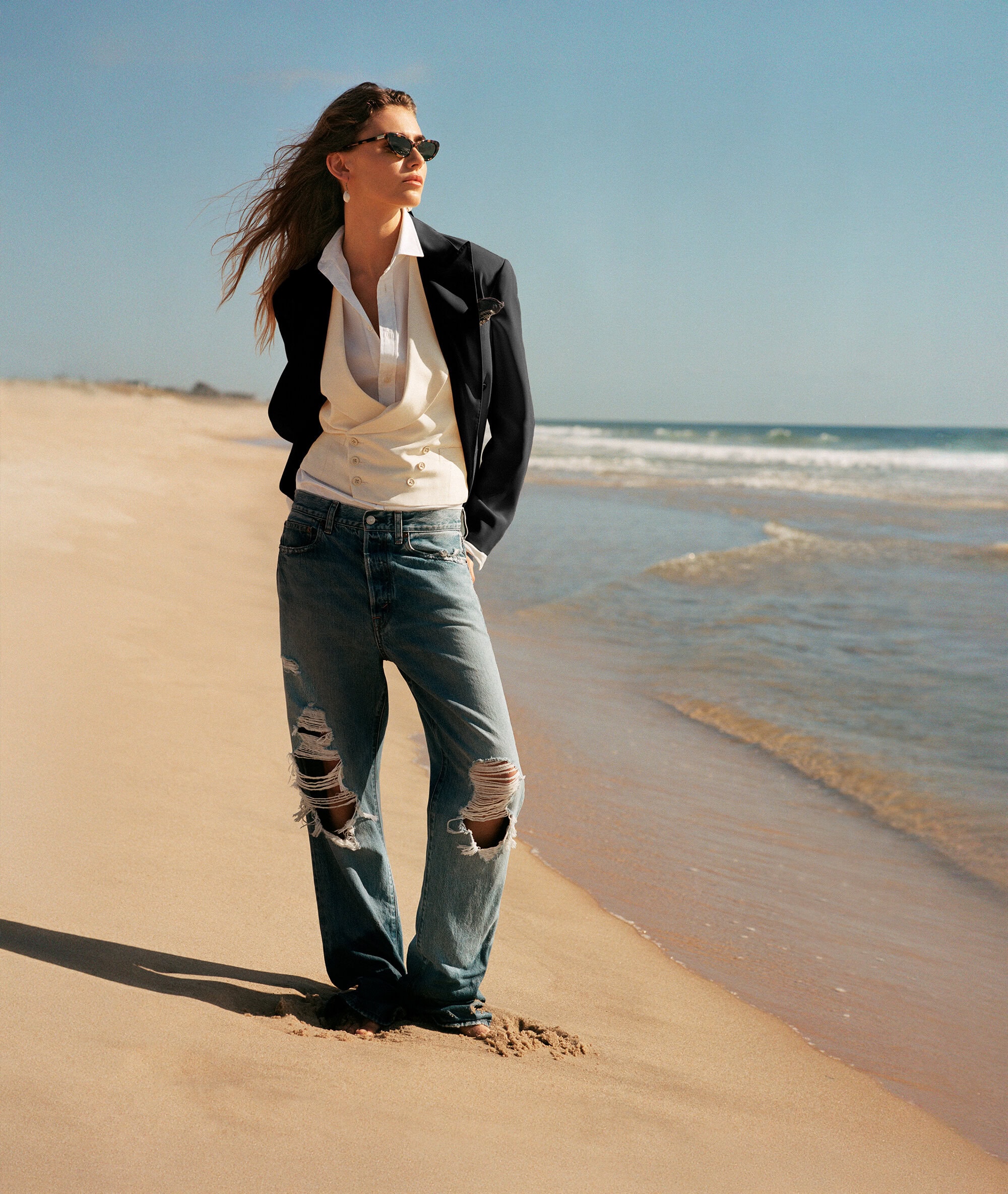 Model wearing a dark blazer, white vest, and distressed denim jeans standing on a sandy beach by the ocean.
