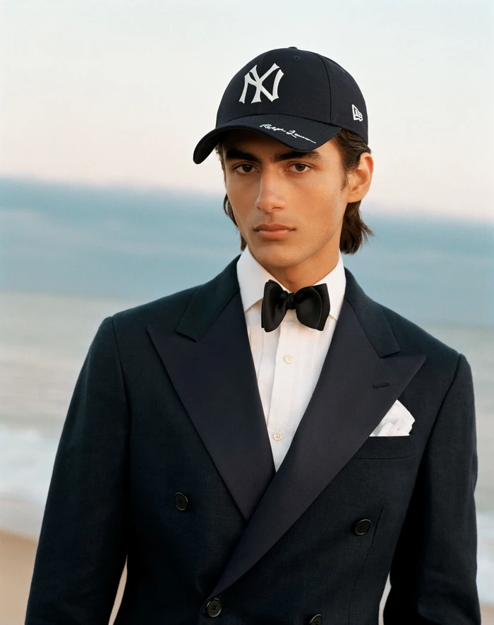 Young man wearing a dark tuxedo jacket, white shirt, black bow tie, and a baseball cap against a blurred coastal backdrop