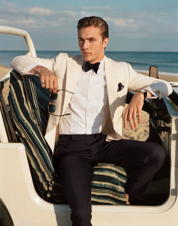 Man wearing ivory suit jacket and bow tie sitting in a vintage beach car with ocean background