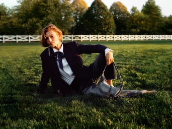 Model seated on green grass wearing a dark blazer, white shirt, and bow tie, holding a handbag, with a white fence behind.