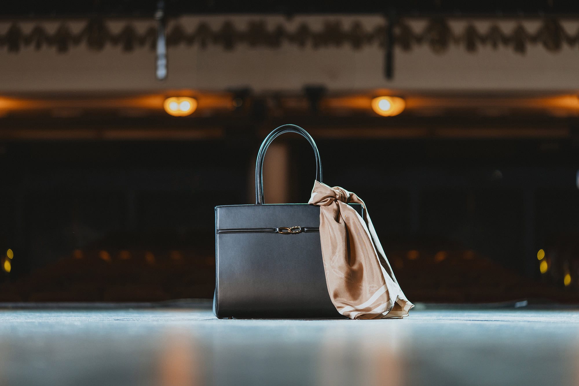Black leather handbag with a tied silk scarf resting on a stage surface with a dimly lit theater auditorium in the background.