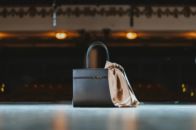 Black leather handbag with a tied silk scarf resting on a stage surface with a dimly lit theater auditorium in the background.