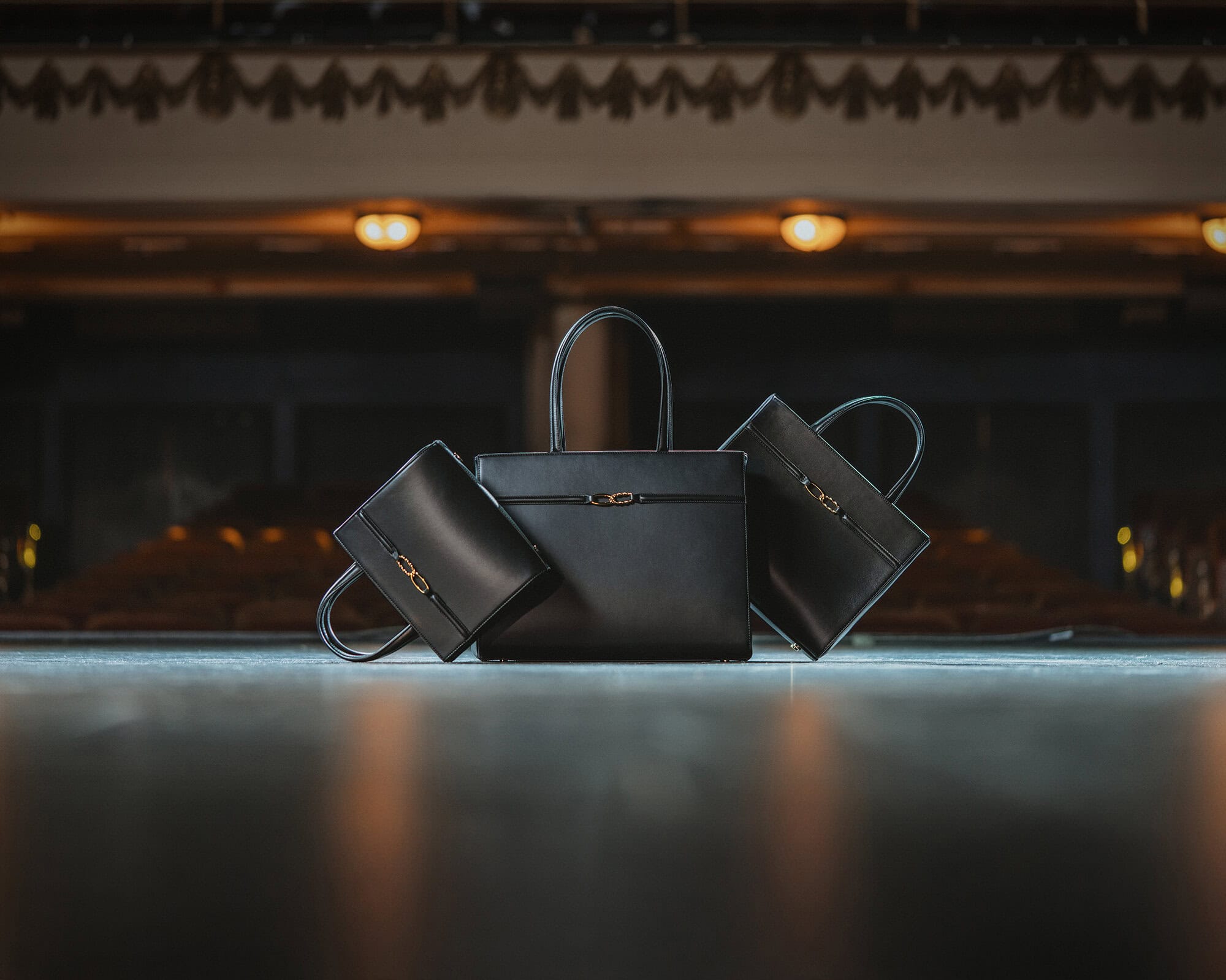 Three black leather handbags displayed centrally on a dark stage with warm overhead lights and a dim auditorium background.