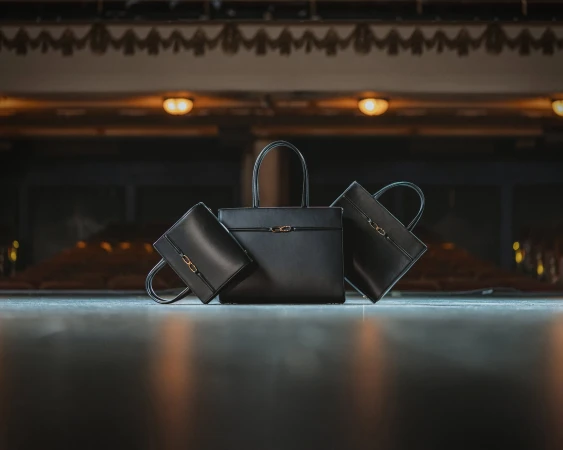 Three black leather handbags displayed centrally on a dark stage with warm overhead lights and a dim auditorium background.